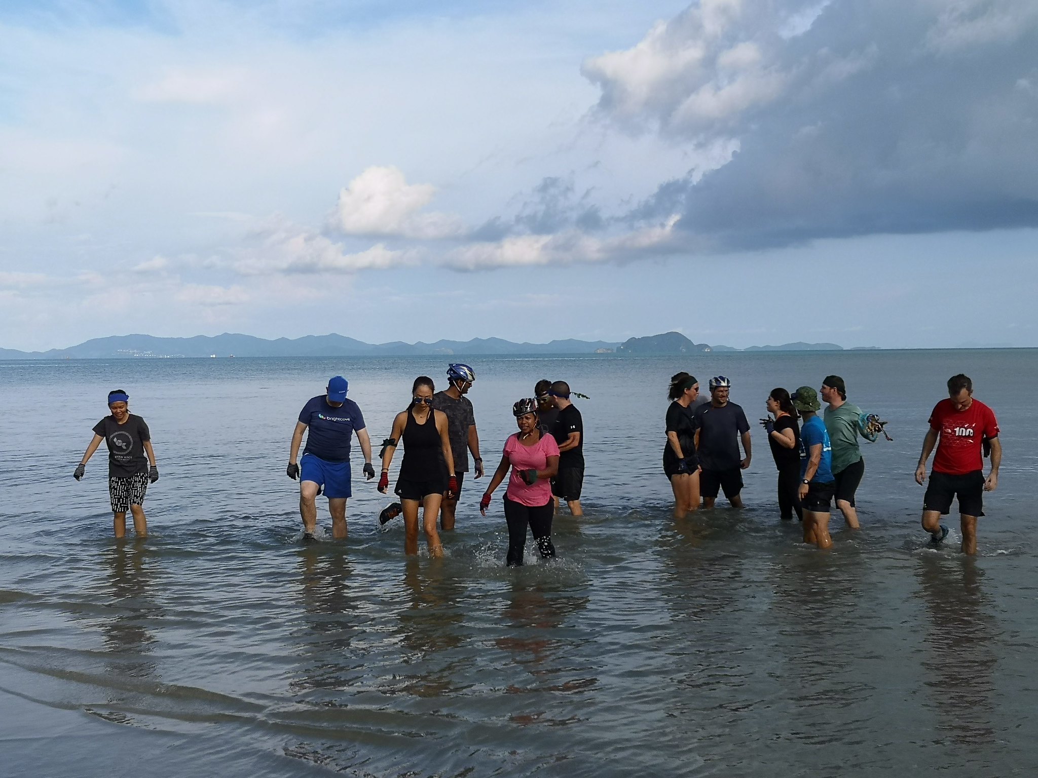 Beaches Hills Island Life A group of team members wading through shallow coastal waters during an outdoor adventure or island excursion, with scenic hills in the background under a cloudy sky.