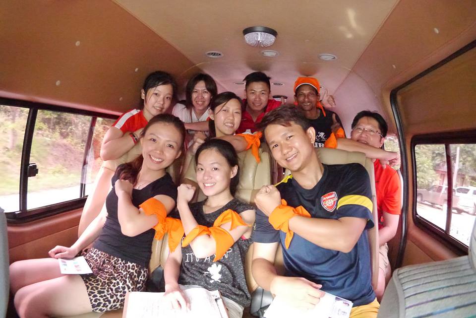 High Value at a Reasonable Cost A cheerful team sitting inside a van, wearing matching orange armbands and posing enthusiastically, representing a cost-effective and fun transportation setup for a team-building event in Thailand.