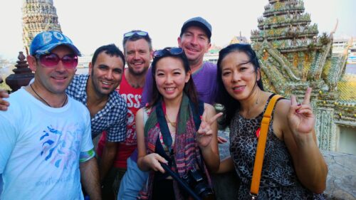 A diverse group of coworkers smiling and posing with peace signs in front of a traditional Thai temple during a cultural team building activity in Bangkok.