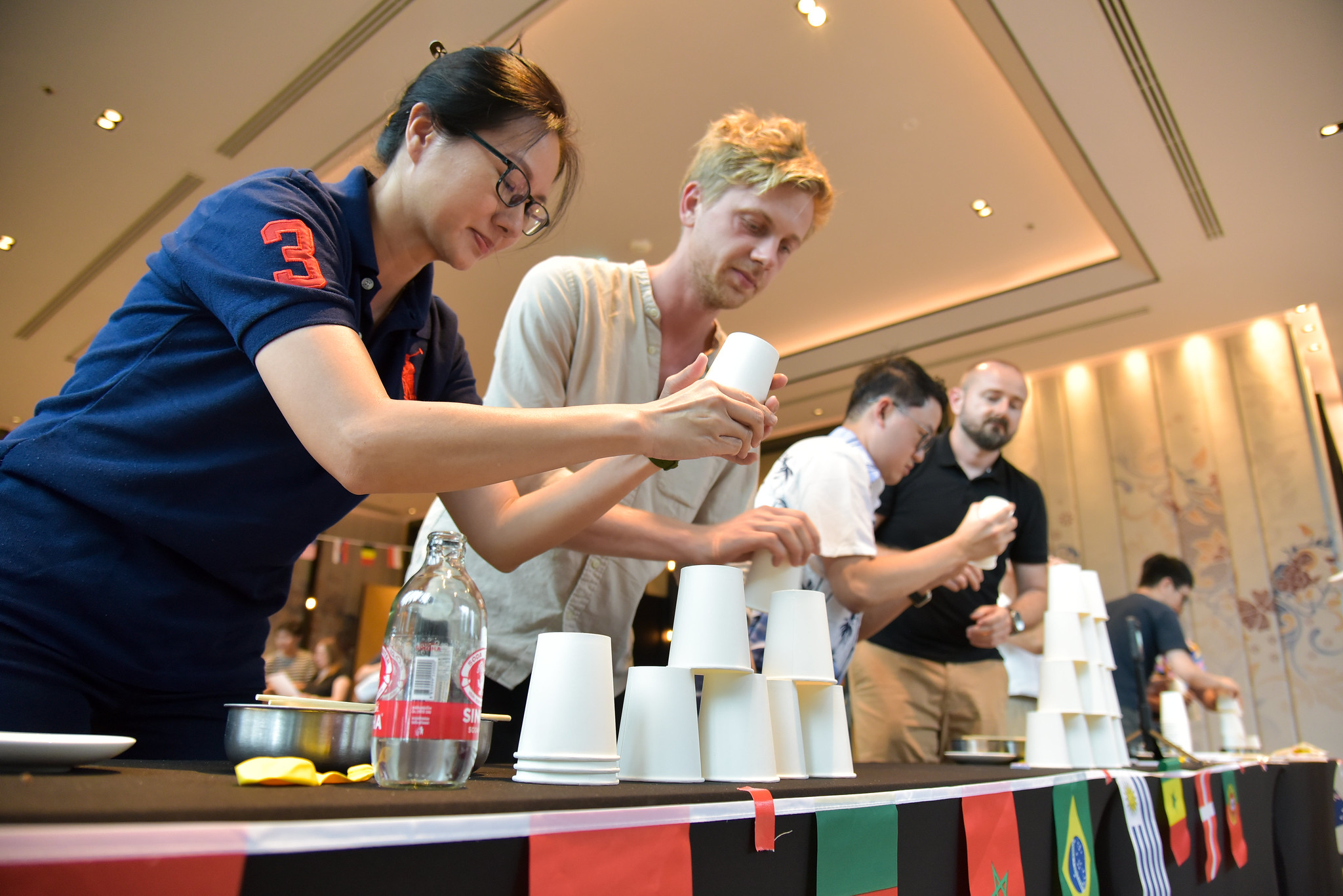 Why Indoor Activities Empower Teams Teams race to build tall stacks of plastic cups on a long table decorated with international flags during a fast-paced indoor team-building activity.