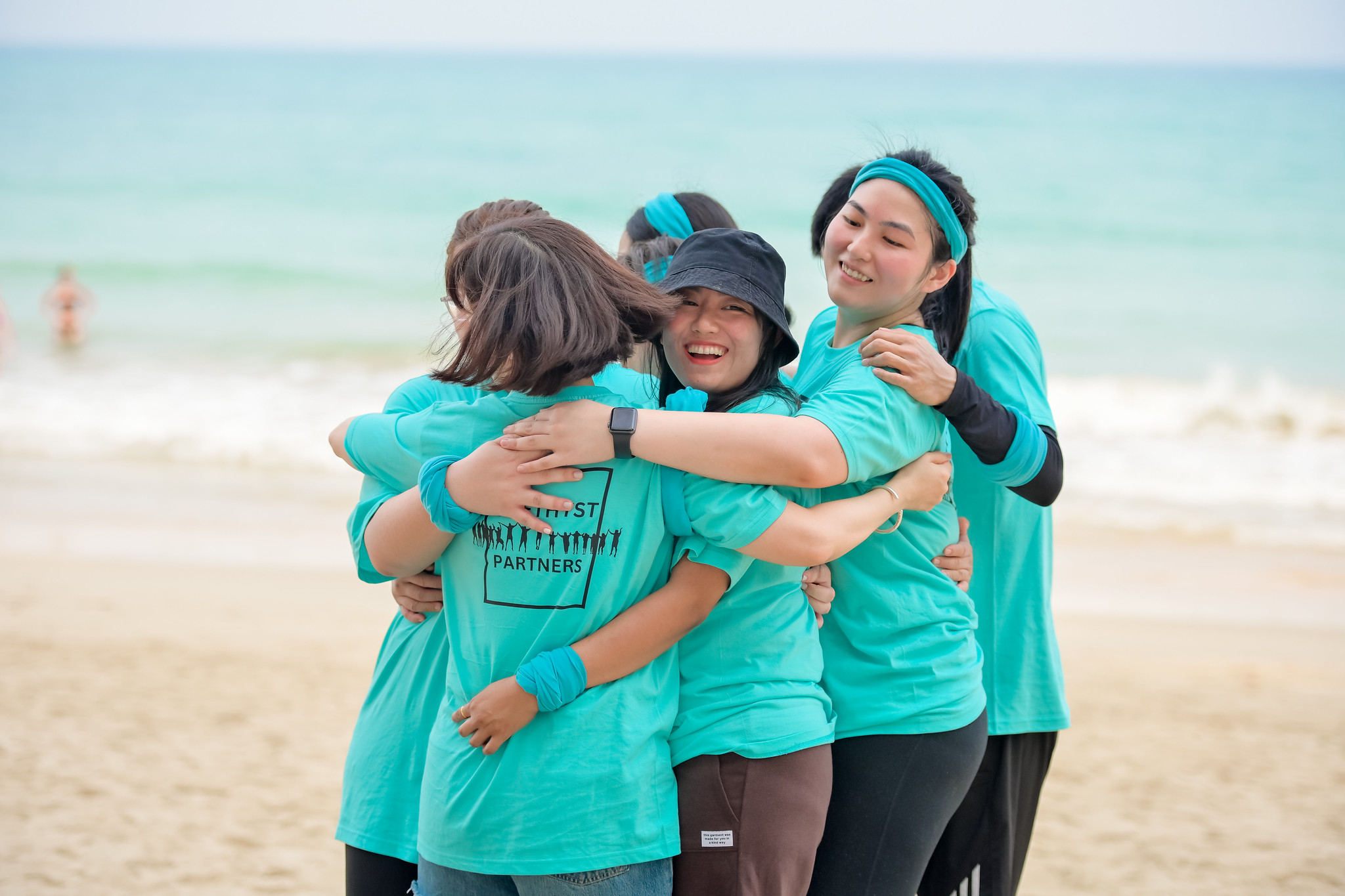 Competitive Relay Missions A group of teammates in turquoise shirts shares a celebratory hug on Phuket beach after completing a competitive relay activity, with the ocean behind them.