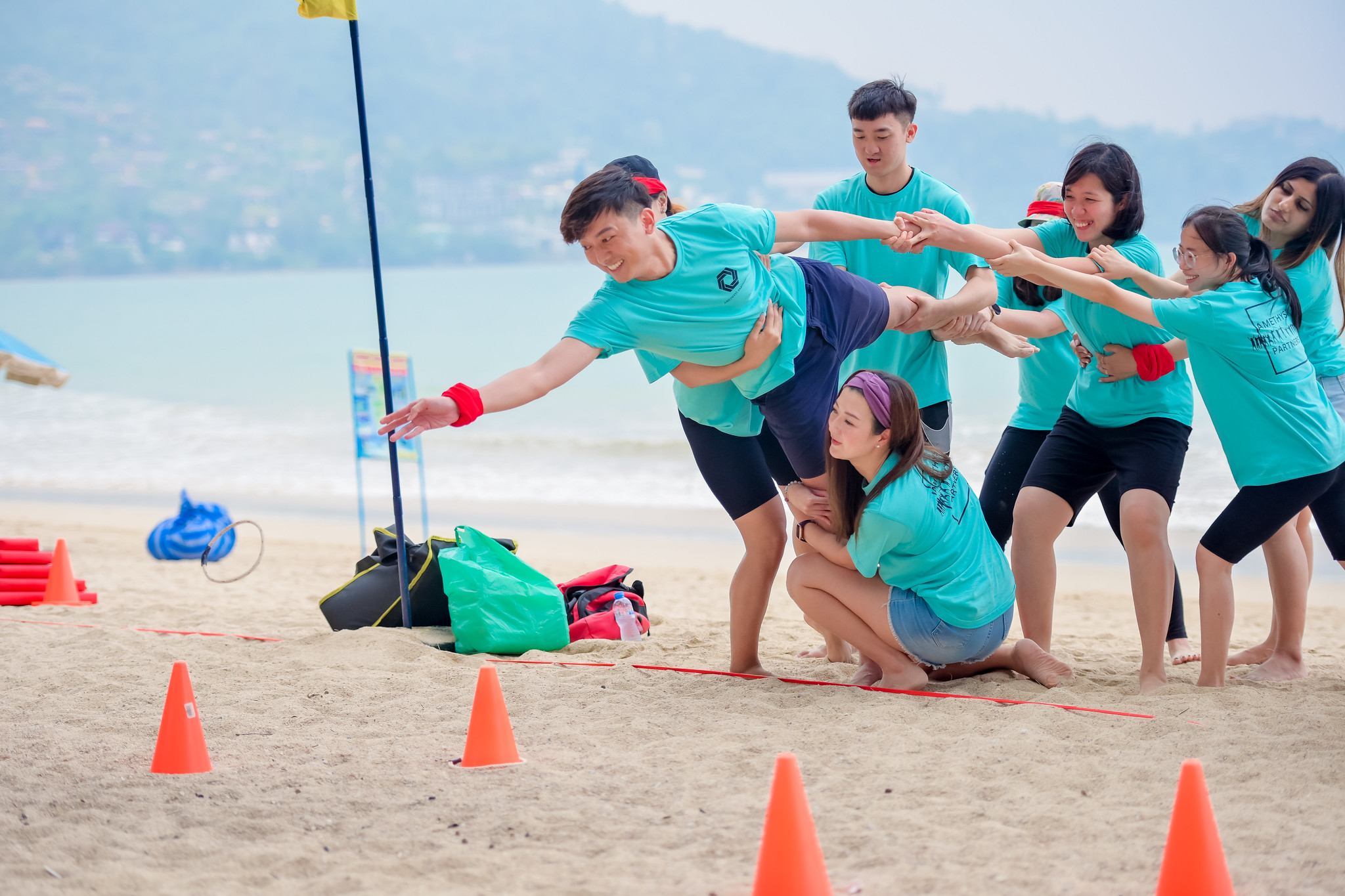 Energizing Icebreaker Games Participants on a Phuket beach engage in a lively icebreaker game, pulling a teammate forward while navigating cones and cheering each other on.